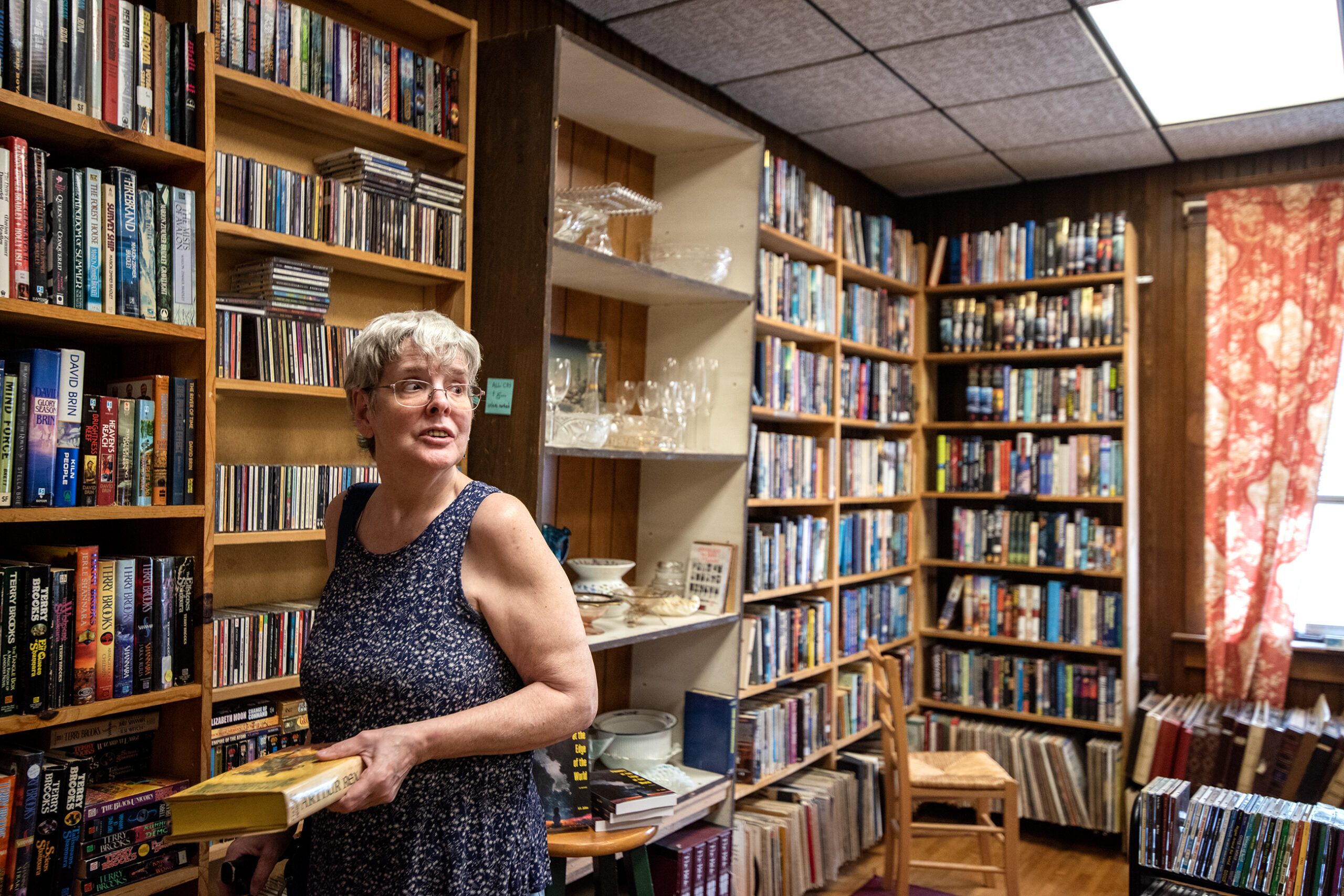 A woman in a sleeveless dress stands holding books in a room filled with shelves of books, CDs, and glassware, with light coming through a window with red curtains.