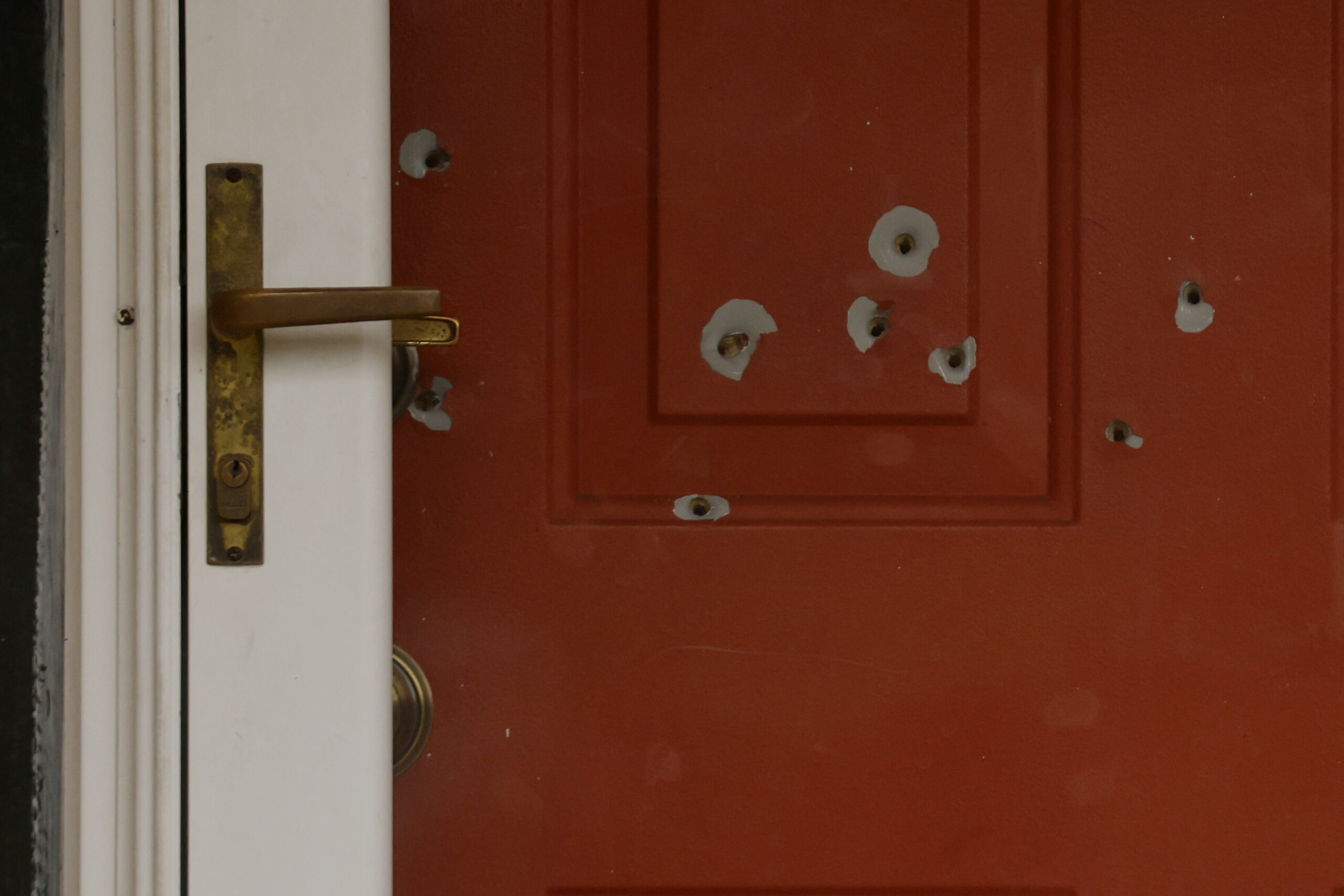 A red door with multiple bullet holes and a brass handle, showing visible damage to the surface.