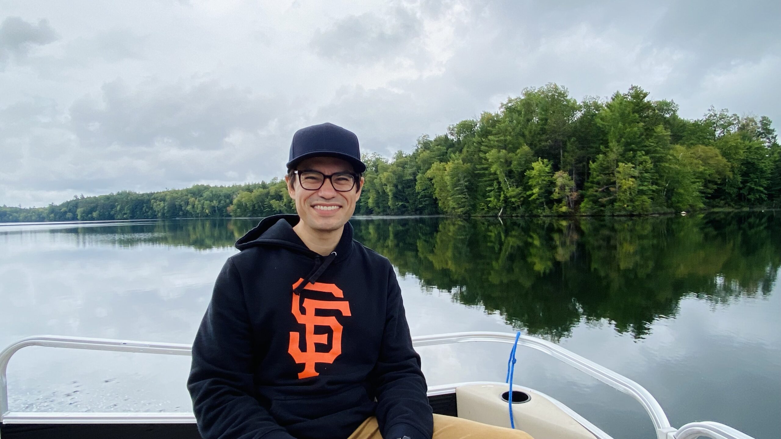 A person wearing a black cap and a black San Francisco Giants hoodie sits on a boat with a calm lake and green trees in the background.