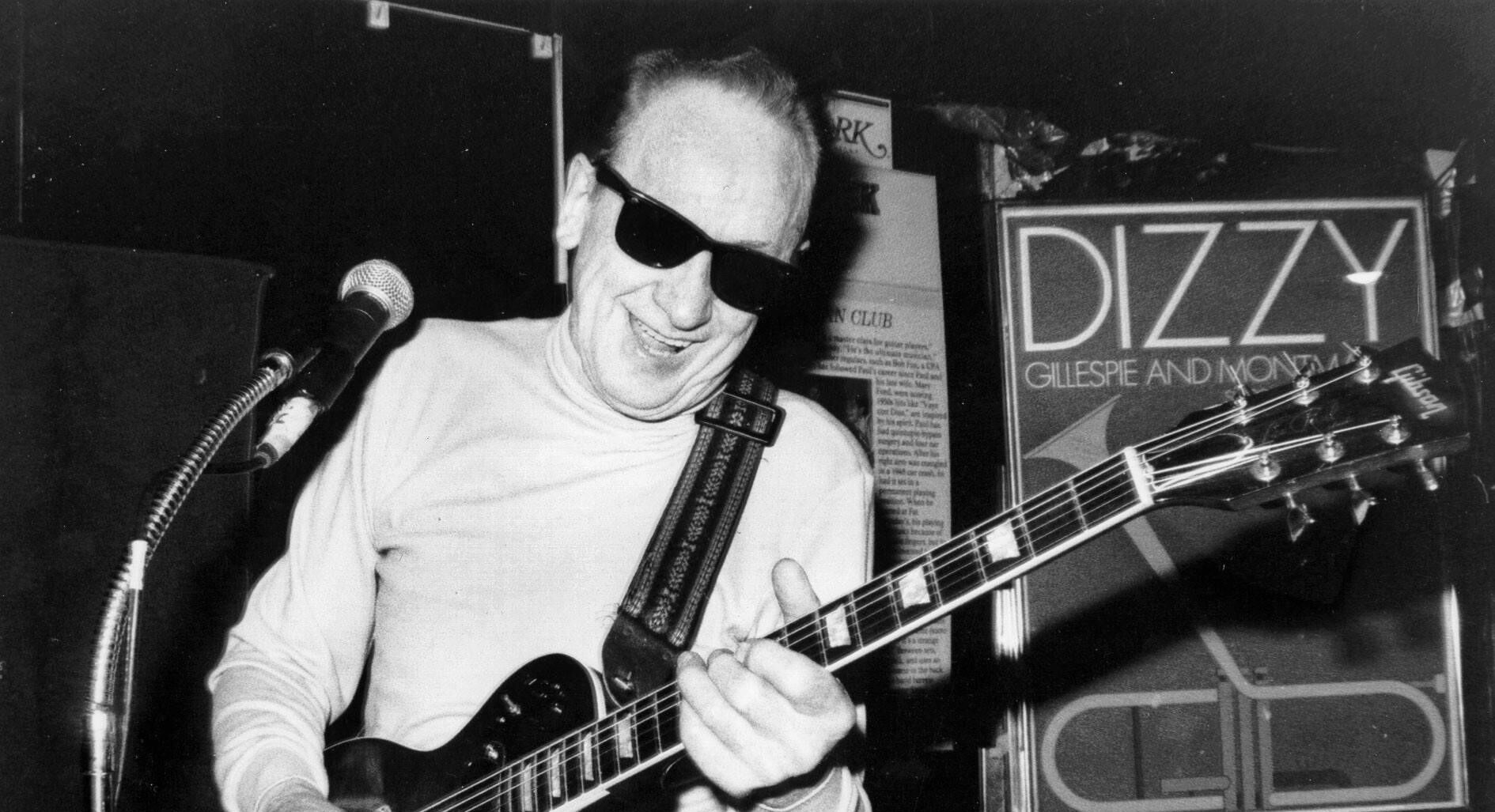 A man wearing sunglasses plays an electric guitar on stage, smiling, with a microphone nearby and a Dizzy Gillespie and Monterey poster in the background.